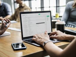 A small team of office workers at a desk, one laptop is open on emails